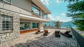 A modern building with a stone facade, a balcony with seating, and a view of a lake and mountains.