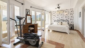 Une chambre à coucher comprenant un lit, du matériel d’exercice, un bureau et de grandes fenêtres.