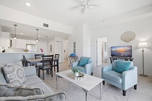 Una sala moderna con un sofá, dos sillones, una mesita de centro y un televisor. Hay un comedor con una mesa y sillas, y una cocina al fondo.