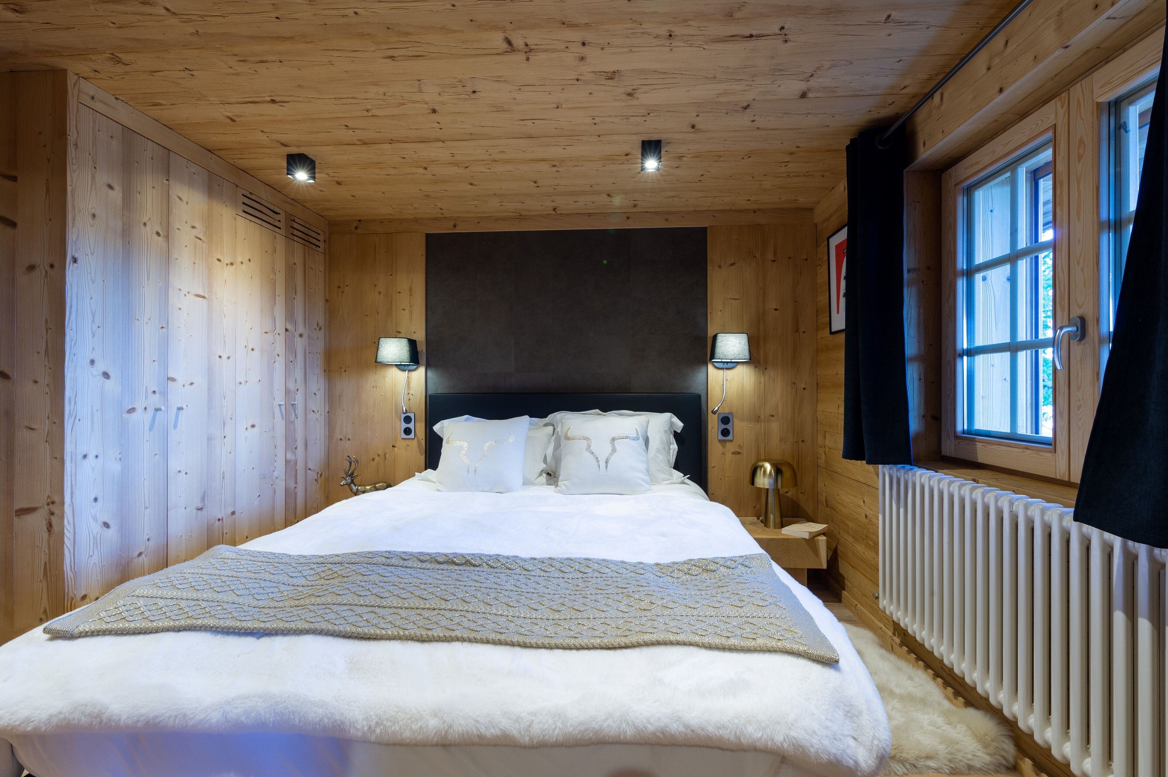 Una habitación con paneles de madera, una cama, dos lámparas fijadas a la pared, una ventana con cortinas y un radiador.