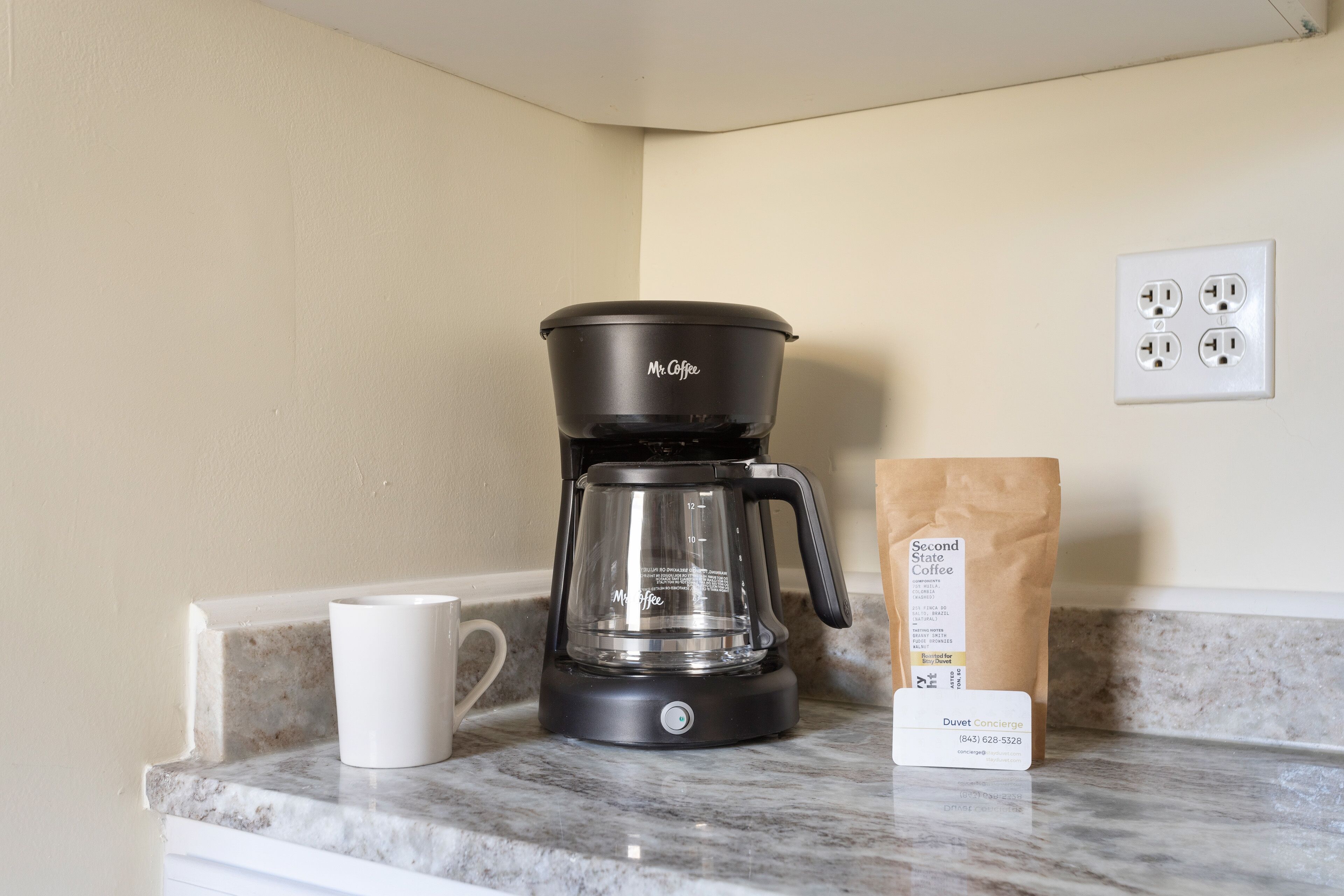 A black Mr. Coffee coffee maker on a marble countertop with a white mug beside it and a brown paper bag with a coffee order label.