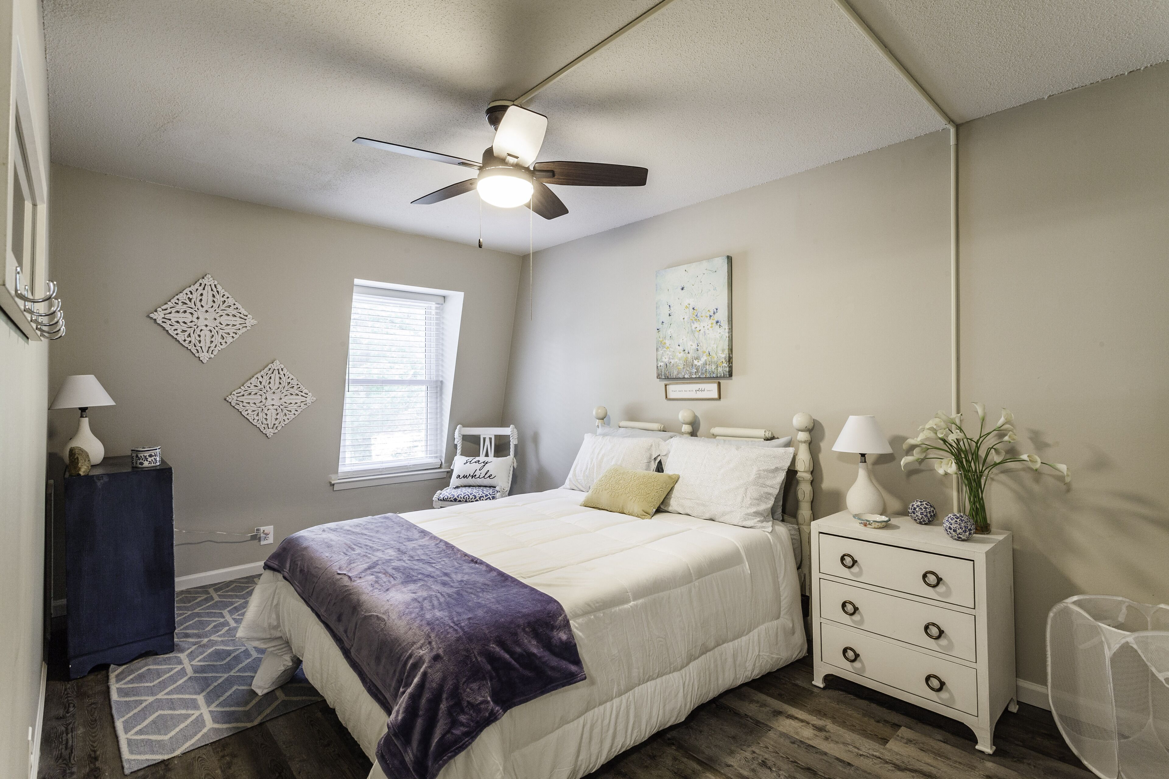 A bedroom with a bed, a nightstand, a ceiling fan, a window, and decorative wall art.