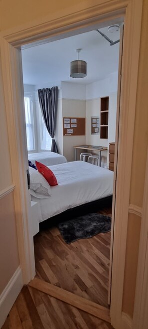Habitación de hotel con cama, escritorio, silla, mesita, tablón de anuncios y ventana con cortinas.