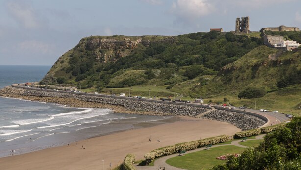 A coastal landscape with a beach, a rocky cliff, and a historic castle on top. - Sally's Berth (Scarborough)