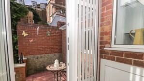 A small patio area with a tiled floor, a white metal gate, a small table with a white teapot and cups, and a brick wall with a window above. - Sally's Berth (Scarborough)
