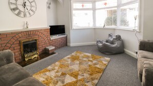 A living room with a fireplace, a large clock, a television, and a bay window with blinds. - Sally's Berth (Scarborough)