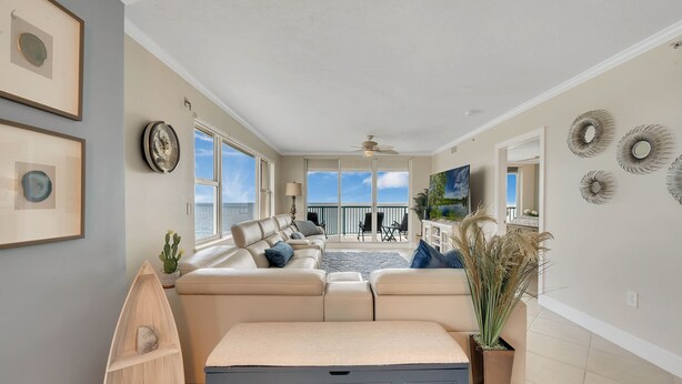 A spacious living room with a beige sofa, a wooden boat shelf, and a large window offering a view of the ocean. - Oceanfront Penthouse ~ Professionally Designed (Daytona Beach Shore)