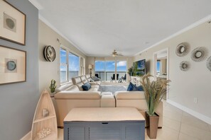 Un salon spacieux avec un canapé beige, une étagère en forme de bateau en bois et une grande fenêtre donnant sur l’océan.