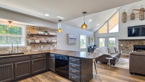 A spacious kitchen with dark cabinetry, a built-in dishwasher, and a large island. There is a stone fireplace, a comfortable seating area with a sofa and a chair, and a large window providing natural light.