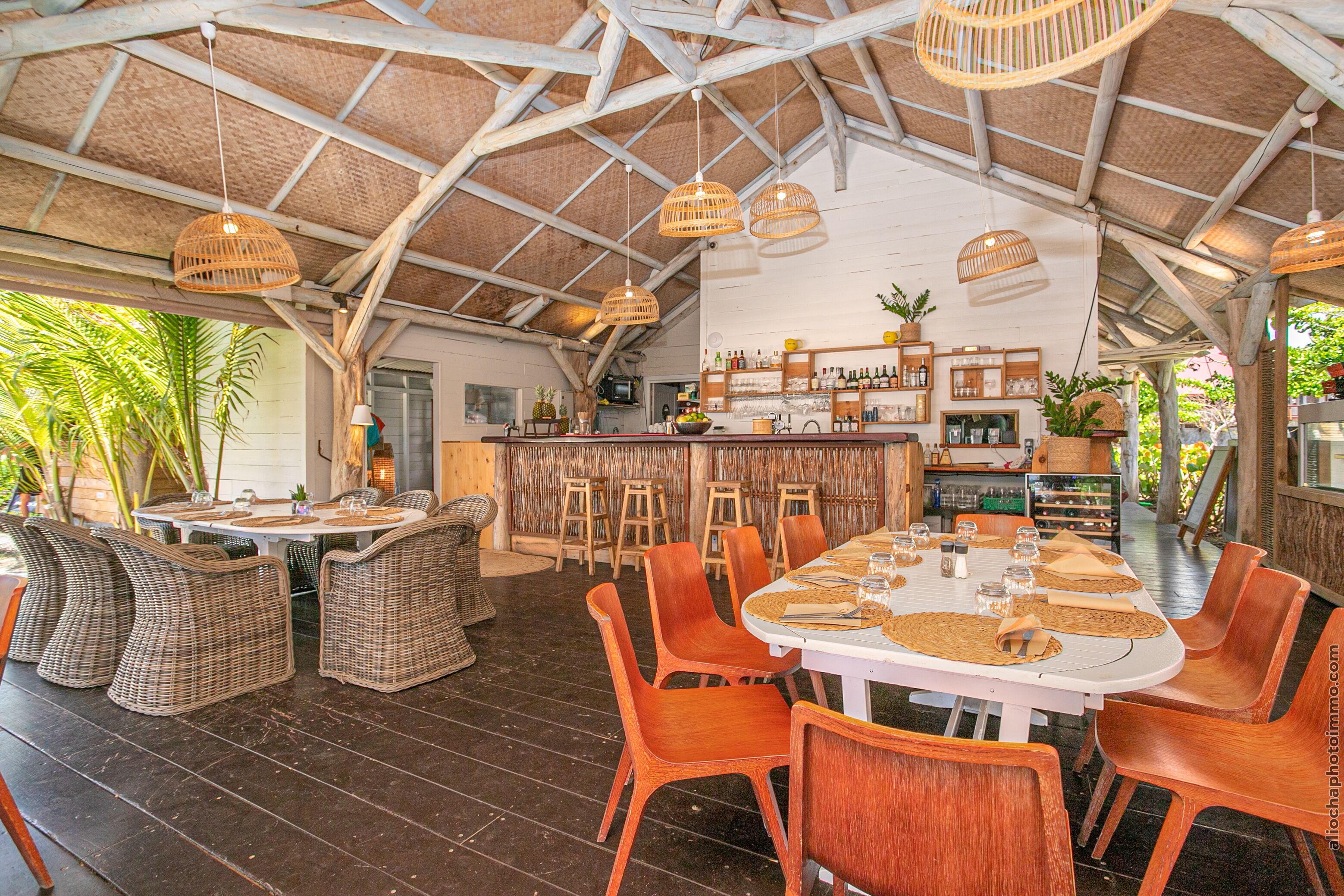 Un espace repas à aire ouverte, doté d’un haut plafond et baigné de lumière naturelle, avec des meubles en osier et un bar central.