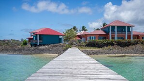 A wooden pier leading to a beachfront property with two buildings, one with a red roof and the other with a blue roof, overlooking clear turquoise water. - Les Villas du Lagon (Le Francois)