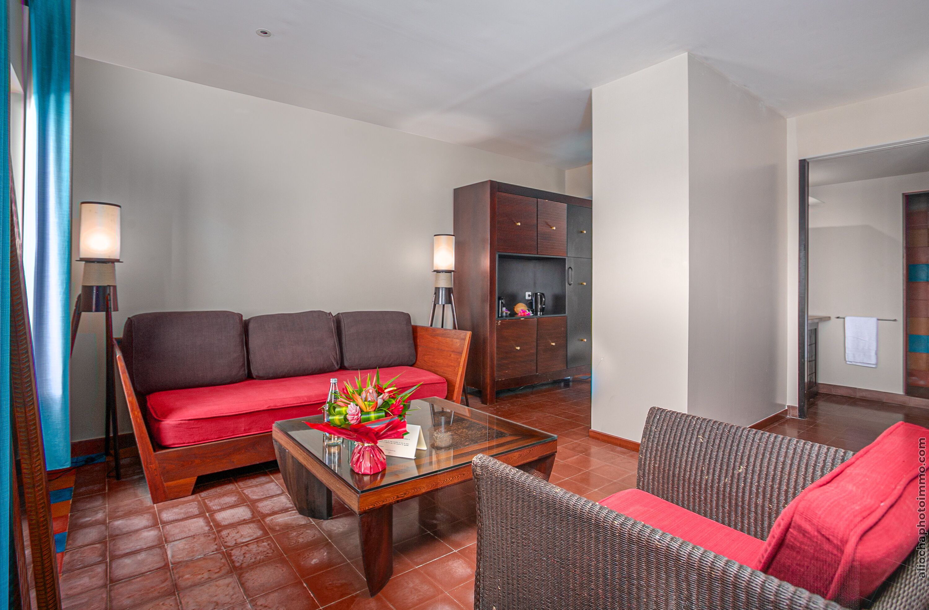 A hotel room with a sofa, a glass coffee table, a wicker chair, a cabinet, and a lamp.