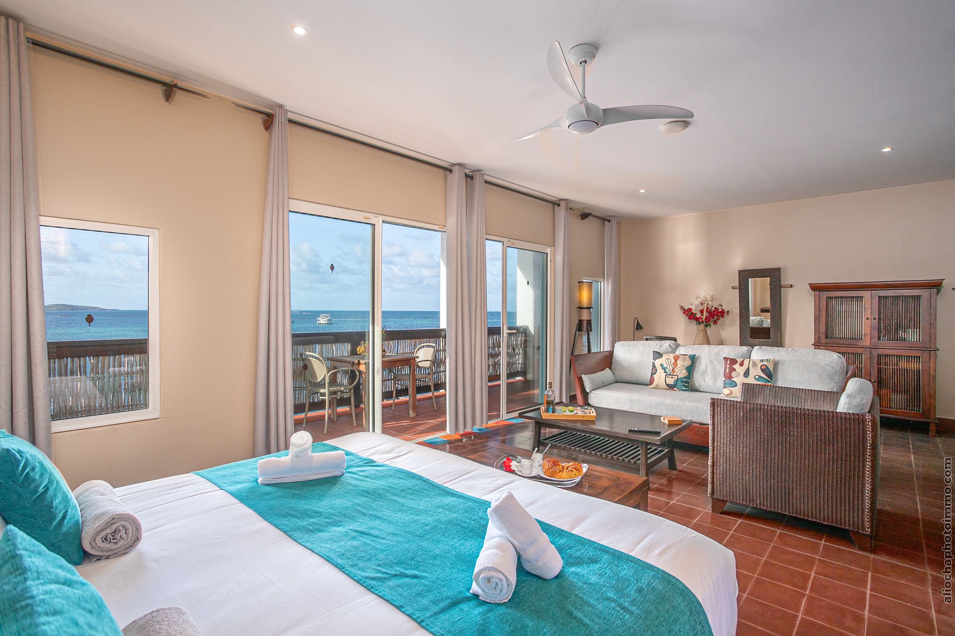 A spacious room with a balcony overlooking the sea, a bed with towels, a sofa, and a dining area.
