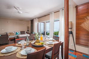 Un comedor con una mesa dispuesta para cuatro, un ventilador de techo y vistas al mar a través de amplios ventanales.
