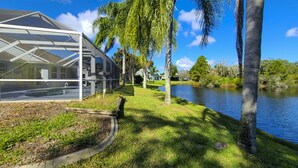 Una moderna estructura de invernadero de vidrio junto a un lago tranquilo, rodeada de exuberante vegetación y palmeras.