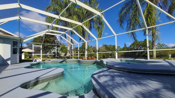 Zona de piscina con cerramiento de vidrio, sillas de descanso y vistas a palmeras y un lago.
