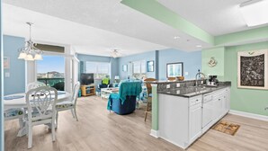 A spacious living area with a dining table and chairs, a kitchen with a central island, and a view through large windows. - Laguna Keyes 612 - Corner Condo - 1 Bedroom - Ocean Front (North Myrtle Beach)