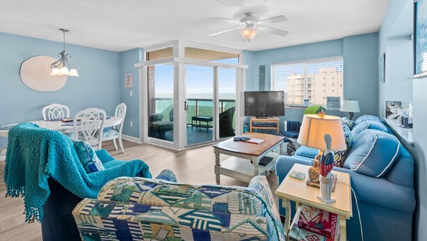 A living room with a blue couch, a coffee table, and a TV. There is a balcony with a view of the ocean. - Laguna Keyes 612 - Corner Condo - 1 Bedroom - Ocean Front (North Myrtle Beach)