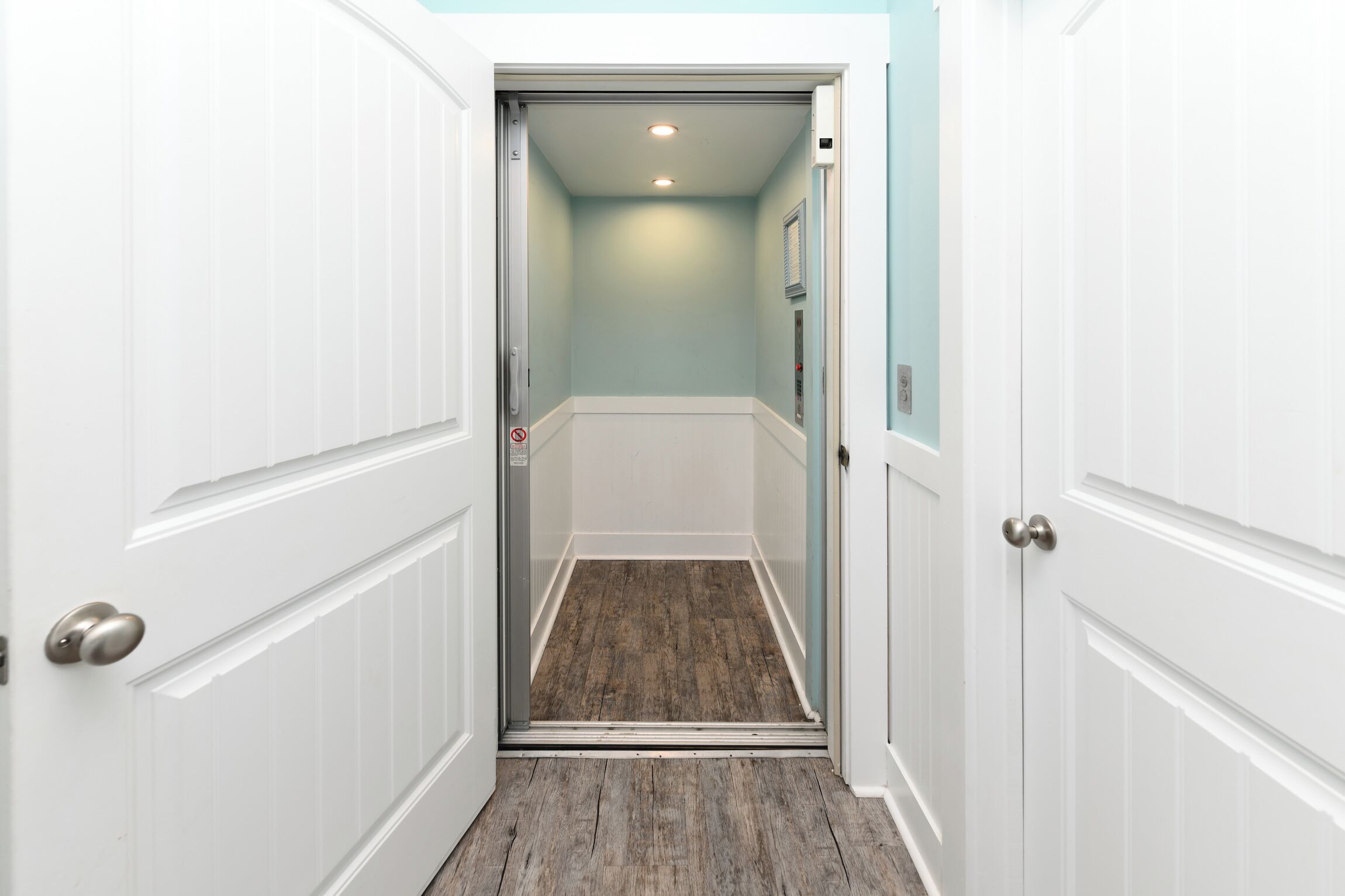 A narrow hallway with white doors and a wooden floor, leading to a room with a light blue wall and a ceiling light.