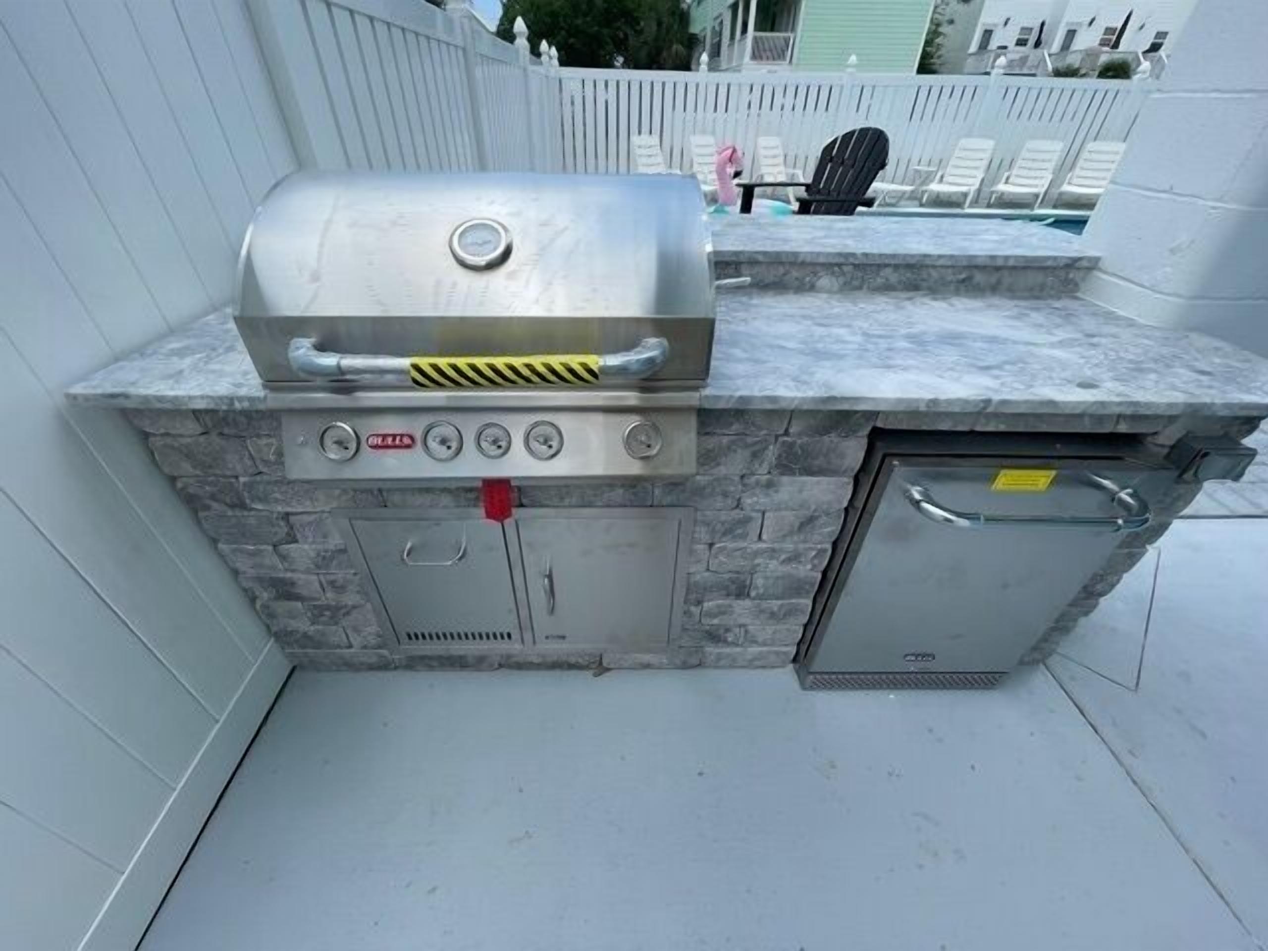 Un barbecue extérieur avec une base en pierre et un couvercle en métal, un petit réfrigérateur et un banc à l’arrière-plan.