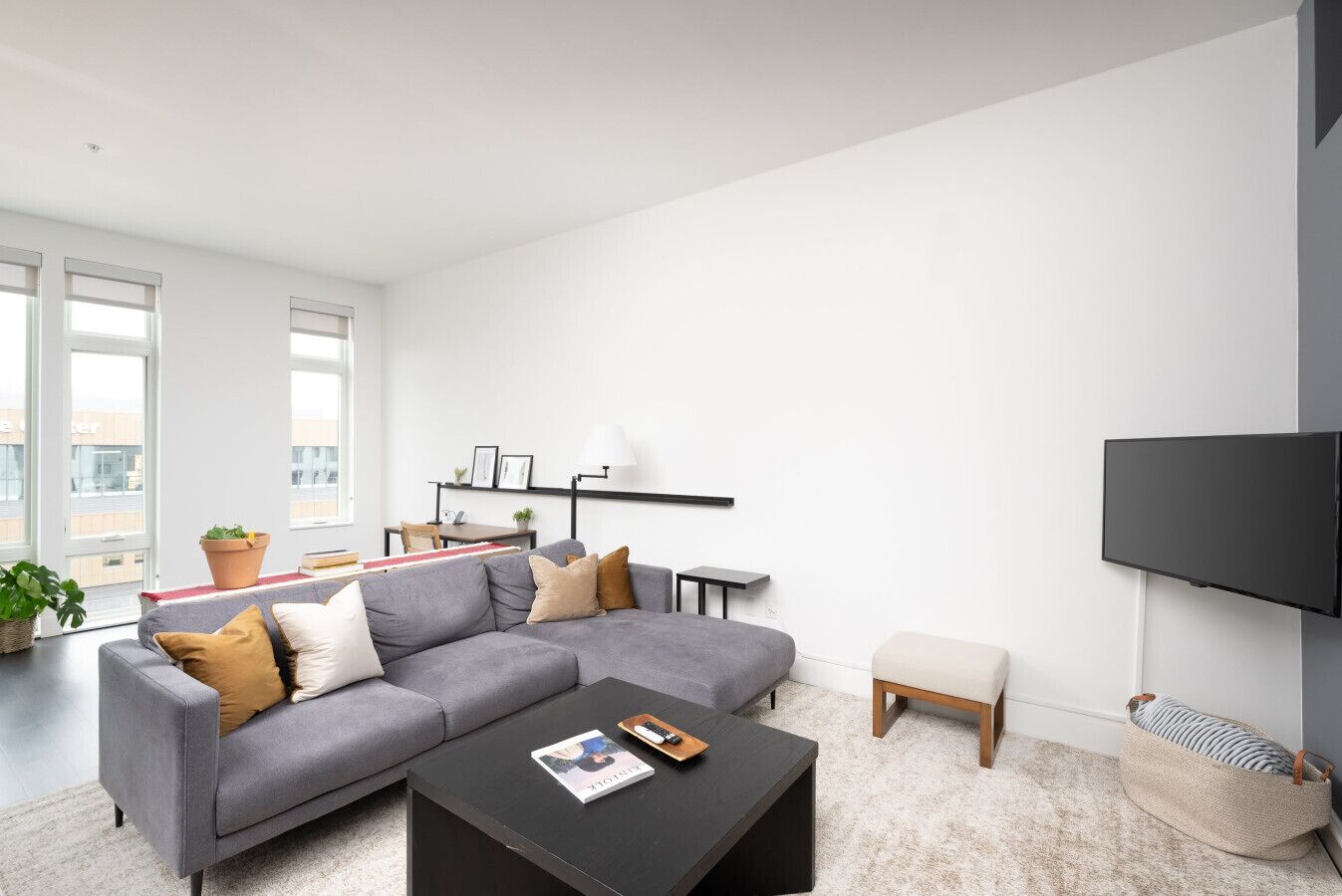 Un salon moderne avec un canapé d’angle gris, une table basse noire, une petite table d’appoint en bois, un fauteuil blanc et un téléviseur fixé au mur.