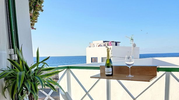 Ein Balkon mit Tisch, einer Champagnerflasche und einem Glas. Ein Blumentopf und ein weißer Stuhl sind ebenfalls vorhanden. Im Hintergrund ist Meer und ein Gebäude zu sehen.
