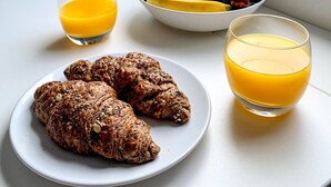 Frühstücksszenario mit Croissants, einer Obstschale und zwei Gläsern Orangensaft auf einem Tisch vor einem Fenster mit Blick auf einen Strand.