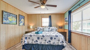 Un dormitorio con paneles de madera, un ventilador de techo, una cama con colcha de temática playera, dos cuadros y una ventana con persianas.