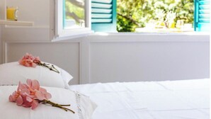 Quarto com roupa de cama branca, janela com persianas azuis e vista para o verde lá fora.