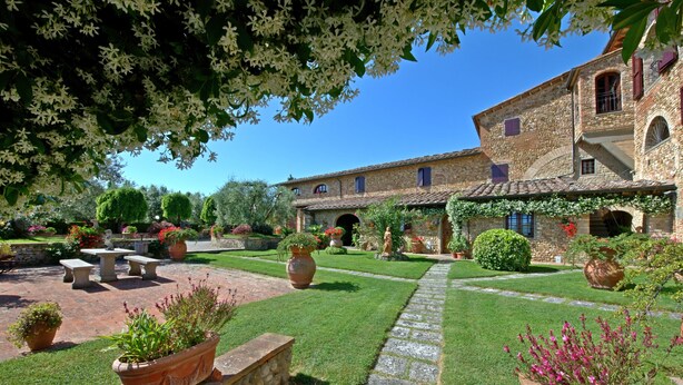 Un patio con un sendero de piedra, plantas en macetas y un edificio con tejado de tejas y ventanas arqueadas.