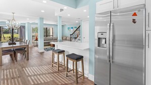 A modern kitchen with a stainless steel refrigerator, wooden bar stools, and a dining area with a chandelier. - "Pipers Perch" - Book Fall Break with Golf Cart! (Miramar Beach)