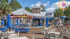 An outdoor dining area with wooden tables and chairs, umbrellas, and a building with a metal roof and wooden accents. - "Pipers Perch" - Book Fall Break with Golf Cart! (Miramar Beach)