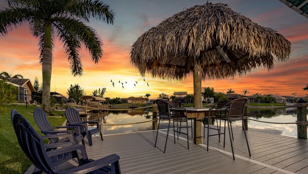 A wooden deck with outdoor furniture, a thatched umbrella, and a body of water reflecting the sunset. - The perfect family vacation home with gulf access/heated pool/tiki bar/ kayaks! (Cape Coral)