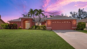 A single-story house with a red garage, a well-maintained lawn, and palm trees. - The perfect family vacation home with gulf access/heated pool/tiki bar/ kayaks! (Cape Coral)