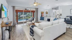 A modern living room with a large sofa, a coffee table, and a TV mounted on the wall. There is a sliding glass door leading to a balcony with a pool view. - The perfect family vacation home with gulf access/heated pool/tiki bar/ kayaks! (Cape Coral)