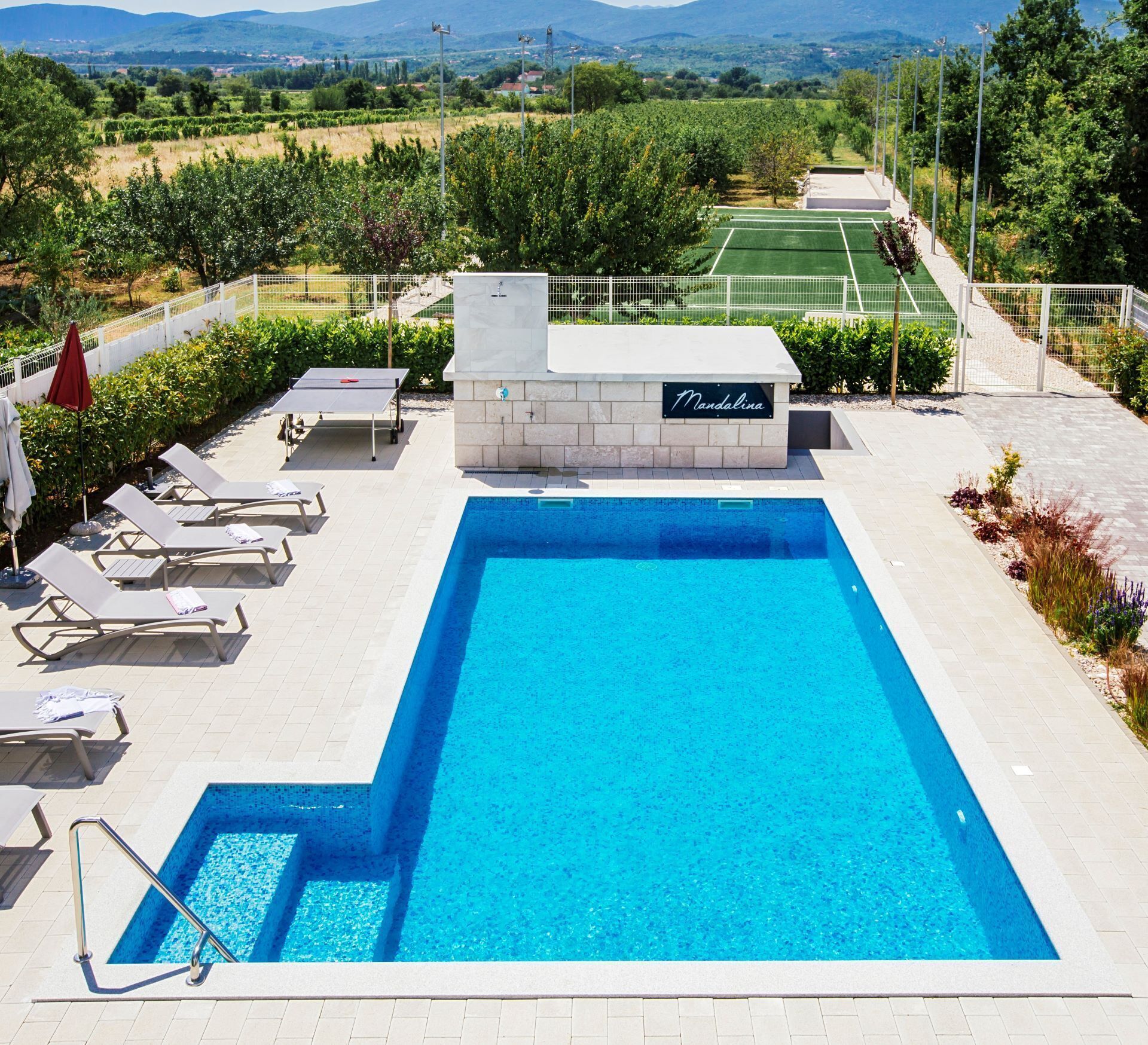 Una piscina rectangular con un letrero que dice "Mandalina", rodeada de sillas de descanso y una cancha de tenis al fondo.