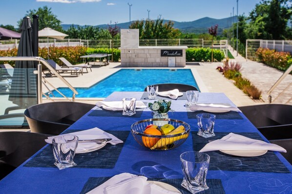 A dining table set with a fruit bowl, glasses, and plates, overlooking a pool and garden. - Luxury Home with Swimming Pool, Professional Tennis Court and Boccia Alley (Donji Proložac)