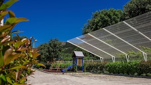 A paved walkway with a playground area, including swings and a slide, surrounded by greenery and a large solar panel structure.
