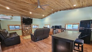 Un salon chaleureux avec une cheminée en pierre, des fauteuils en cuir, une cuisine équipée d’un réfrigérateur et un ventilateur de plafond.