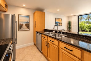 A modern kitchen with a stainless steel refrigerator, a dishwasher, wooden cabinets, a granite countertop, and a window with a view of greenery. - Garden Cottage G5 + Patio | North Coast Village (Oceanside)