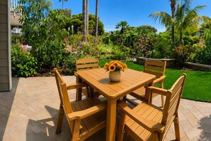 Una mesa de madera al aire libre con sillas, un florero con flores y un jardín bien cuidado con plantas variadas y césped.