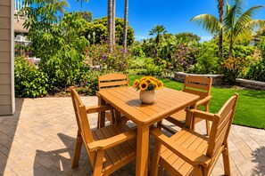 An outdoor wooden table with chairs, a vase of sunflowers, and a garden with various plants and trees.
