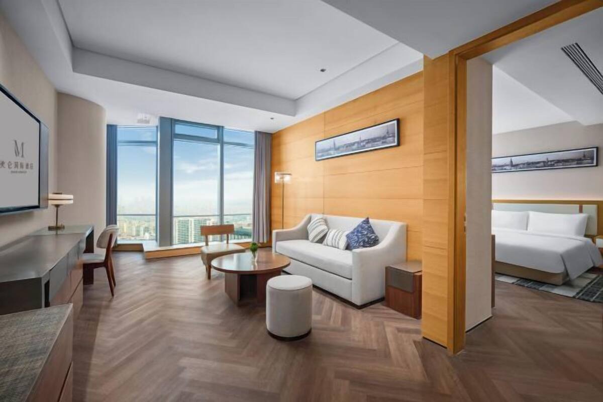 A modern hotel room with a large window, a sofa, a dining table, and a bed.