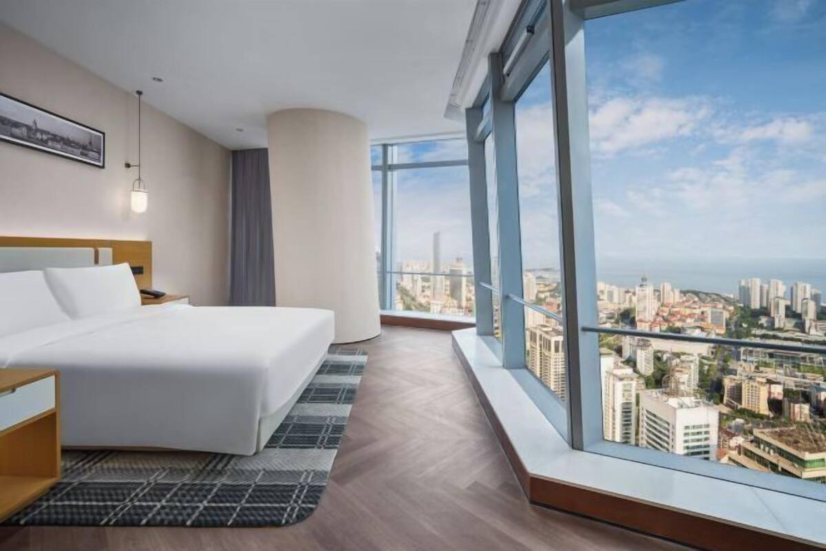 A modern hotel room with a large bed, a nightstand, a desk, and a view of the cityscape through large windows.