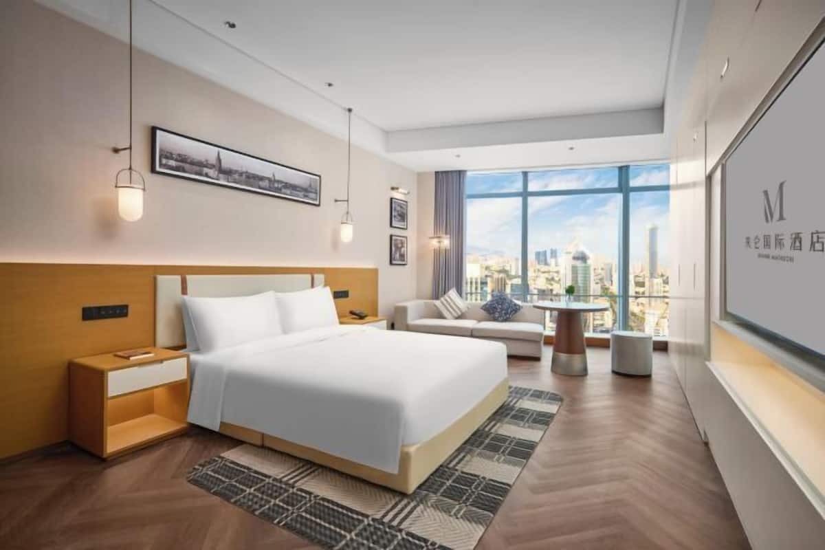 A modern hotel room with a large bed, a sofa, a small table, and a city view through large windows.