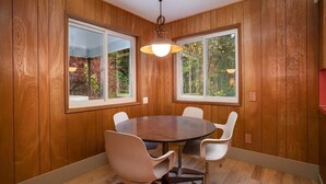 Un coin repas avec une table ronde et quatre chaises, des murs lambrissés en bois et de grandes fenêtres donnant sur la forêt.