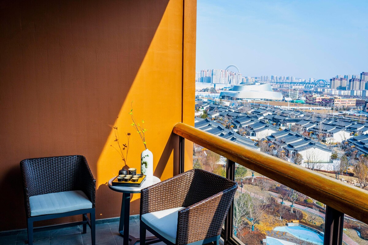 A balcony with wicker chairs, a small table with a vase, and a view of a cityscape with traditional buildings and a modern structure.