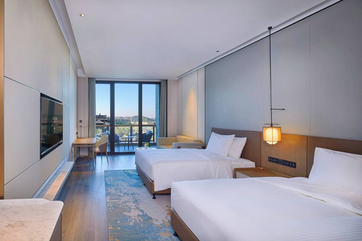 A modern hotel room with two beds, a desk, and a balcony with a view.