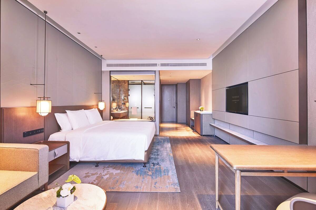 A modern hotel room with a large bed, a TV, a desk, and a bathroom visible through an open door.
