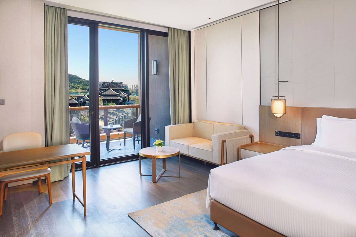 A hotel room with a bed, a sofa, a table, and a chair. There is a view of traditional buildings outside the window.